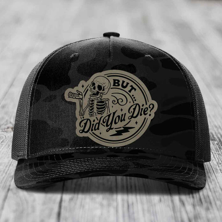 Black camouflage trucker hat with a beige circular patch featuring a skeleton and the text &