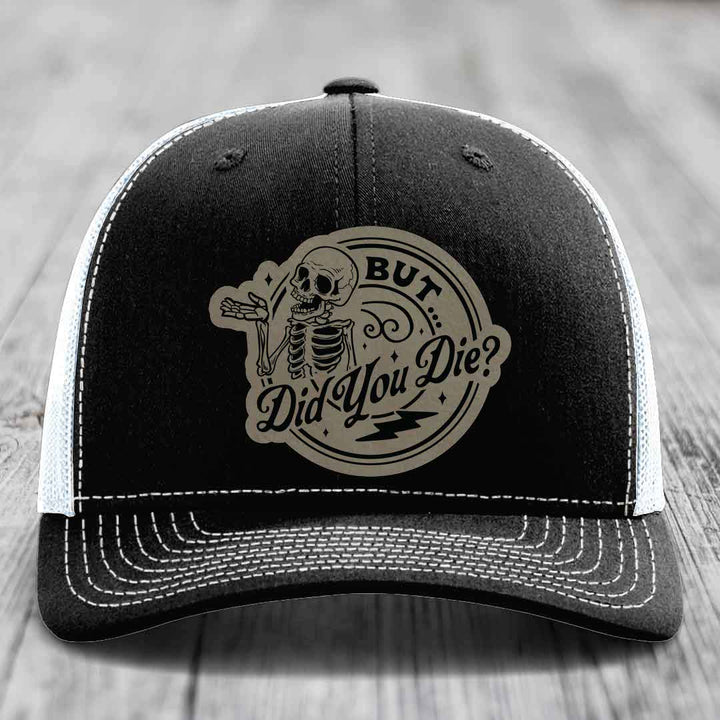 Black trucker hat with white stitching and a beige circular patch featuring a skeleton and the text &