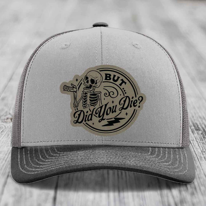 Grey trucker hat with a circular leather patch featuring a skeleton and the text &