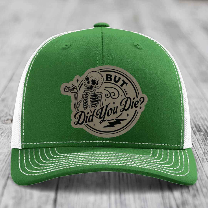 Green trucker hat with white mesh back and stitching, featuring a circular beige patch with a skeleton and text &