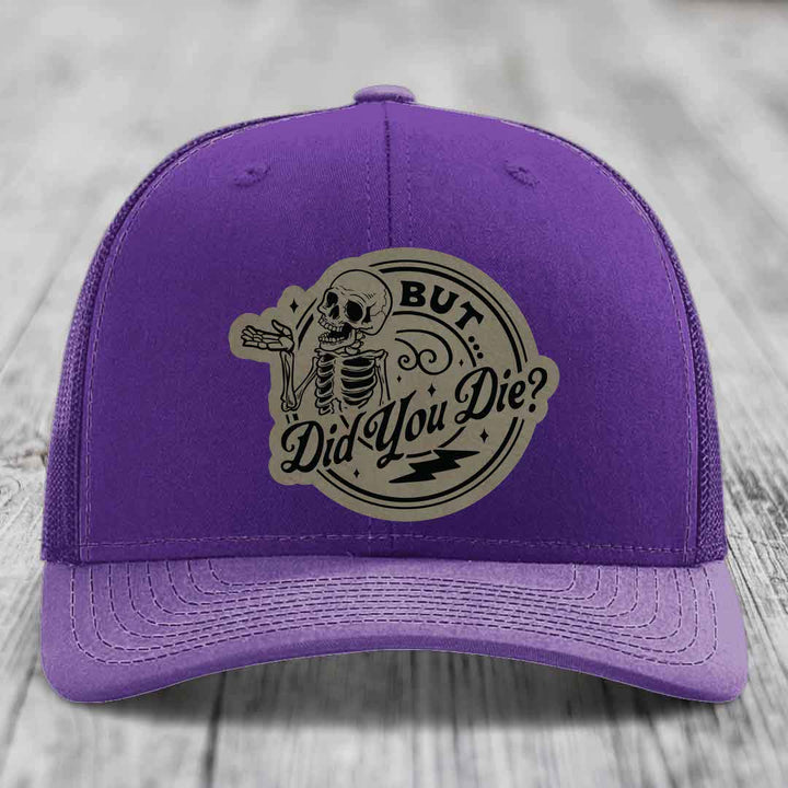 Purple trucker hat with a beige circular patch featuring a skeleton and the text &