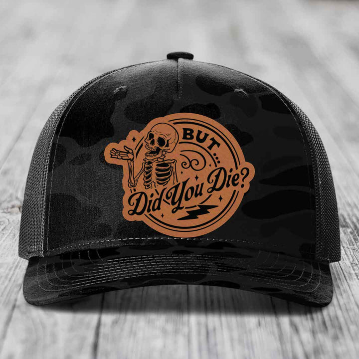 Black camouflage trucker hat with a tan leatherette patch featuring a skeleton and the text &