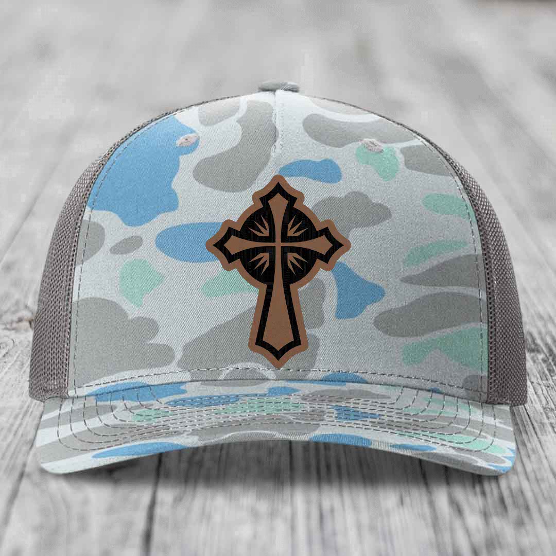 Camouflage trucker hat with brown cross patch on front, gray mesh back, resting on wooden surface.