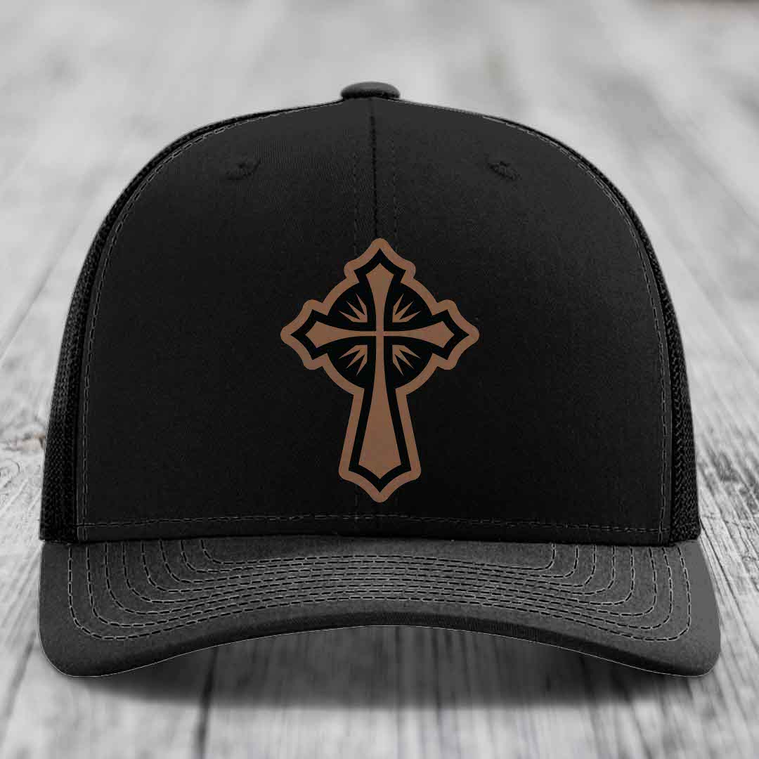 Black trucker hat with brown cross patch on front, sitting on wooden surface.