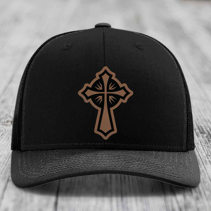 Black trucker hat with brown cross patch on front, sitting on wooden surface.