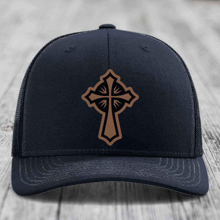 Black trucker hat with brown cross leather patch on front, light gray stitching, wood grain background.