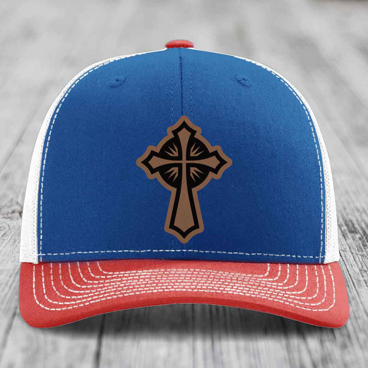 Blue and red trucker hat with a brown leatherette cross patch on the front.