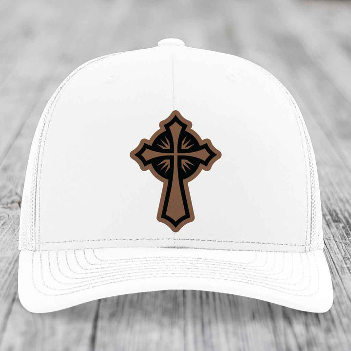 White trucker hat with a brown and black leatherette cross patch on the front.