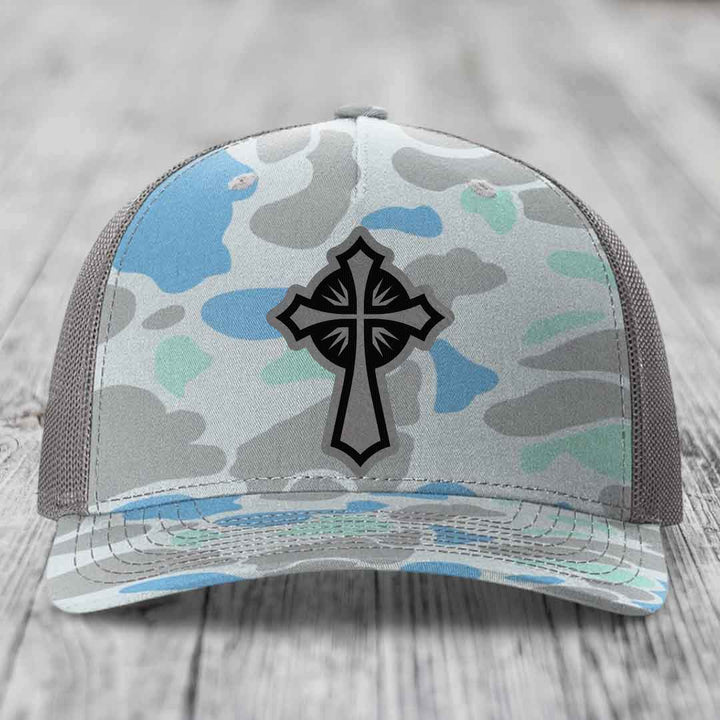 Camouflage trucker hat with black cross patch on front, gray mesh back, sitting on wooden surface.
