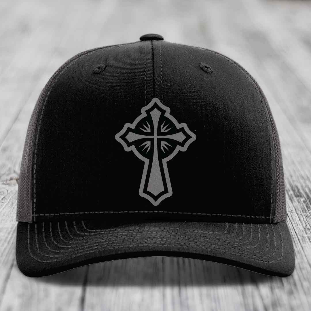 Black trucker hat with grey Celtic cross patch on front, resting on wooden surface.