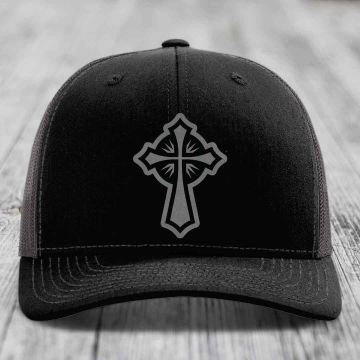 Black trucker hat with grey Celtic cross patch on front, resting on wooden surface.