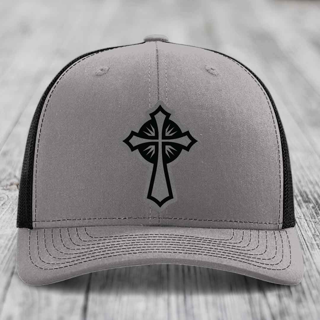 Grey trucker hat with black cross patch on front, black mesh back, white wood background.