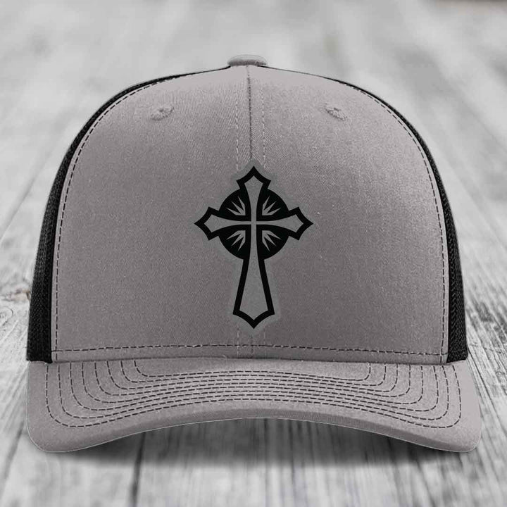 Grey trucker hat with black cross patch on front, black mesh back, white wood background.
