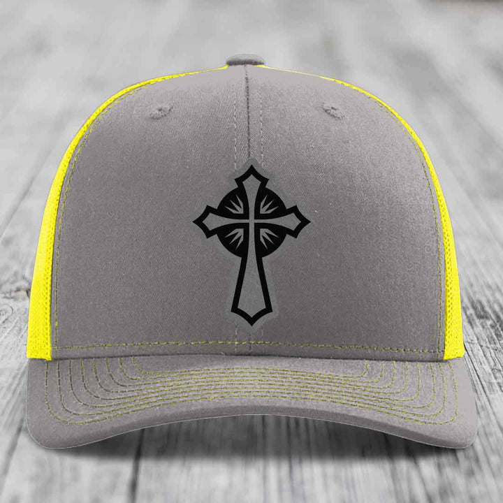 Grey trucker hat with black cross patch and yellow mesh sides and stitching.