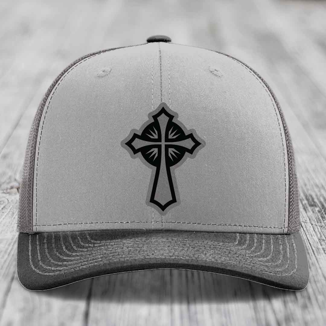 Grey trucker hat with black cross patch on front, placed on wooden surface.