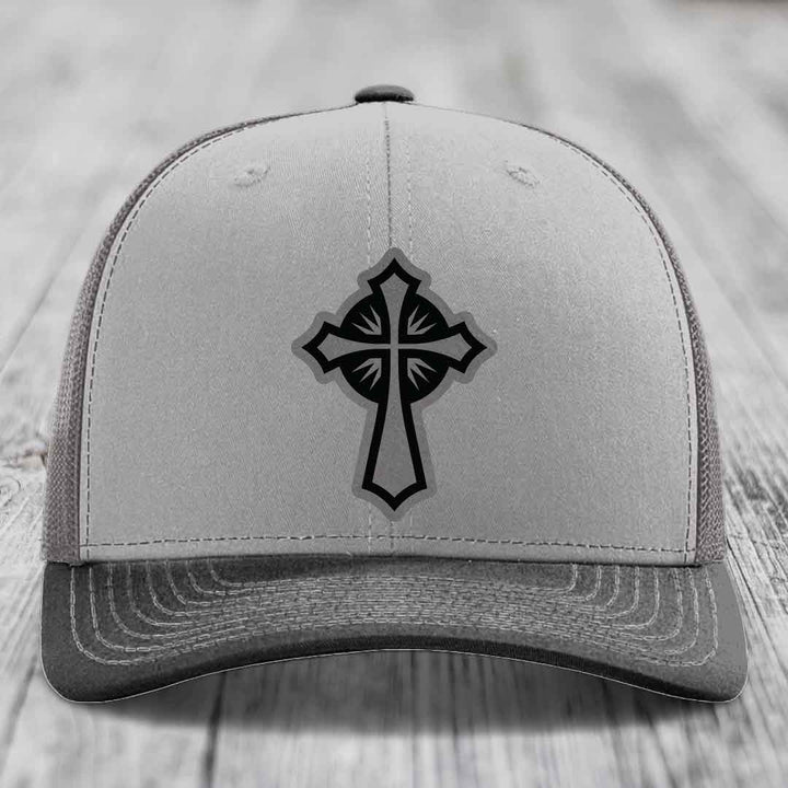 Grey trucker hat with black cross patch on front, placed on wooden surface.