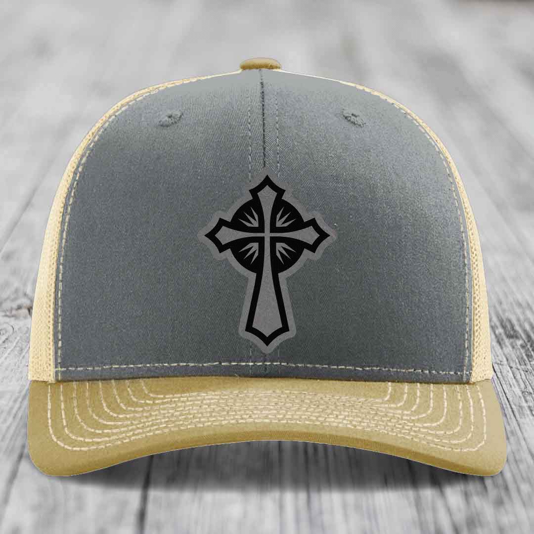 Grey and beige trucker hat with black cross patch on front, white stitching, wooden background.