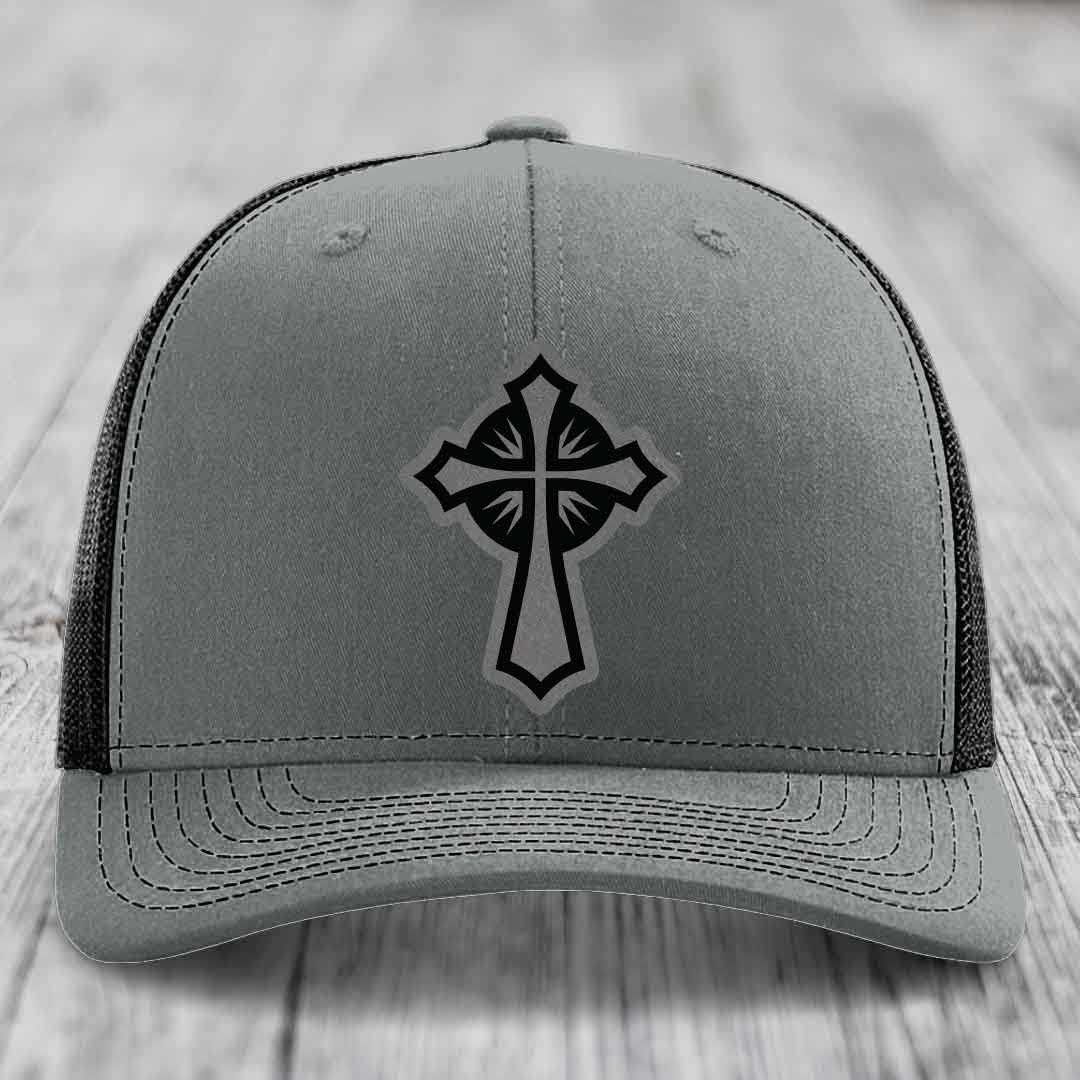 Grey trucker hat with black cross patch on front, black mesh back, sitting on wooden surface.