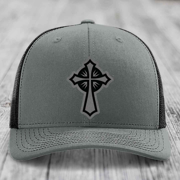 Grey trucker hat with black cross patch on front, black mesh back, sitting on wooden surface.