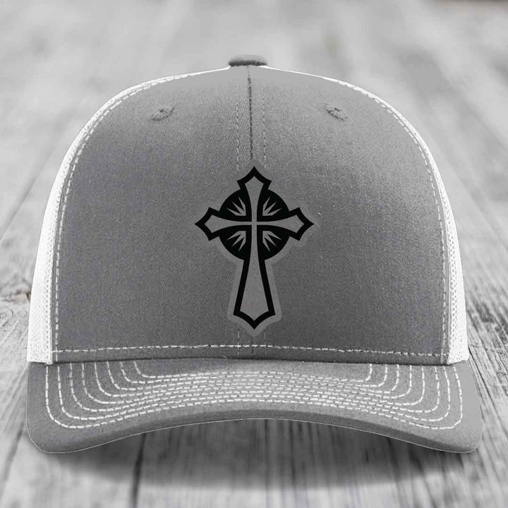 Grey trucker hat with black cross patch on front, white mesh back and white stitching.