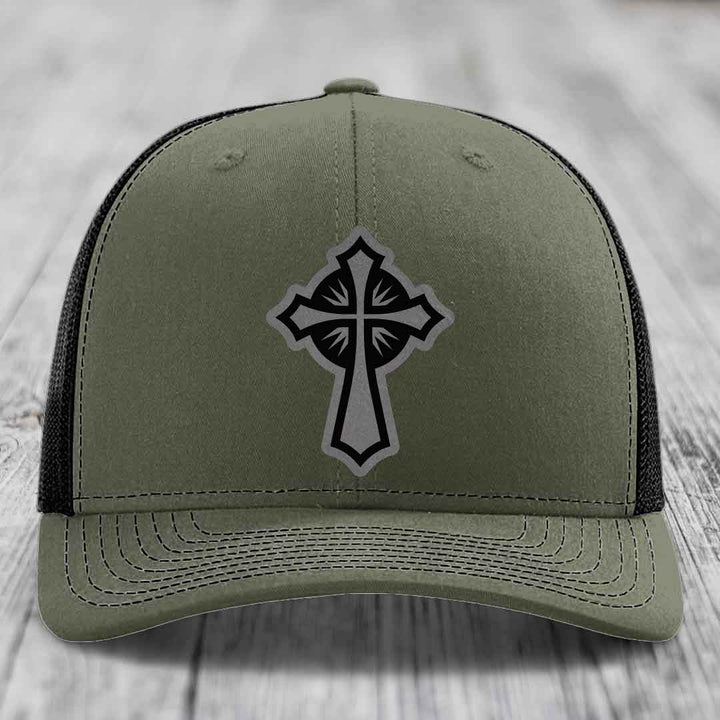 Olive green trucker hat with black cross patch on front, black mesh back.