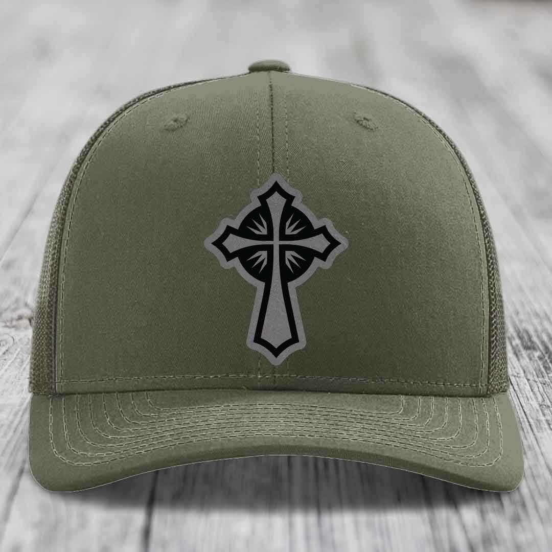 Olive green trucker hat with black cross patch on front, placed on wooden surface.