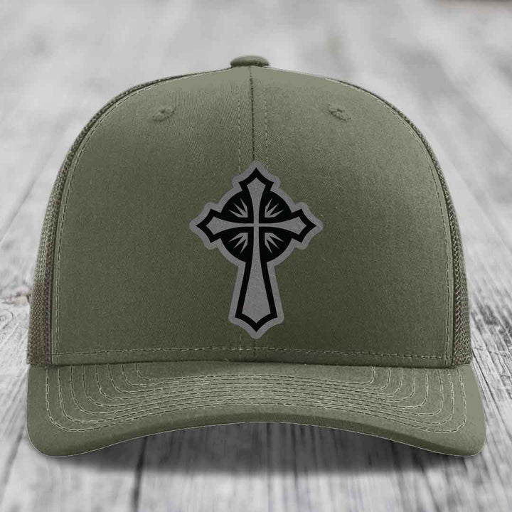 Olive green trucker hat with black cross patch on front, placed on wooden surface.