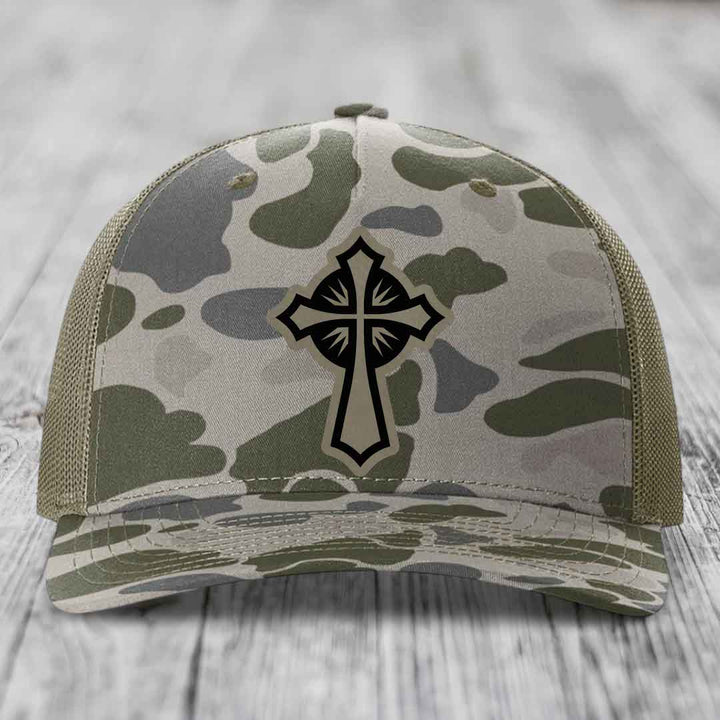 Camouflage trucker hat with black cross patch on front, placed on wooden surface.