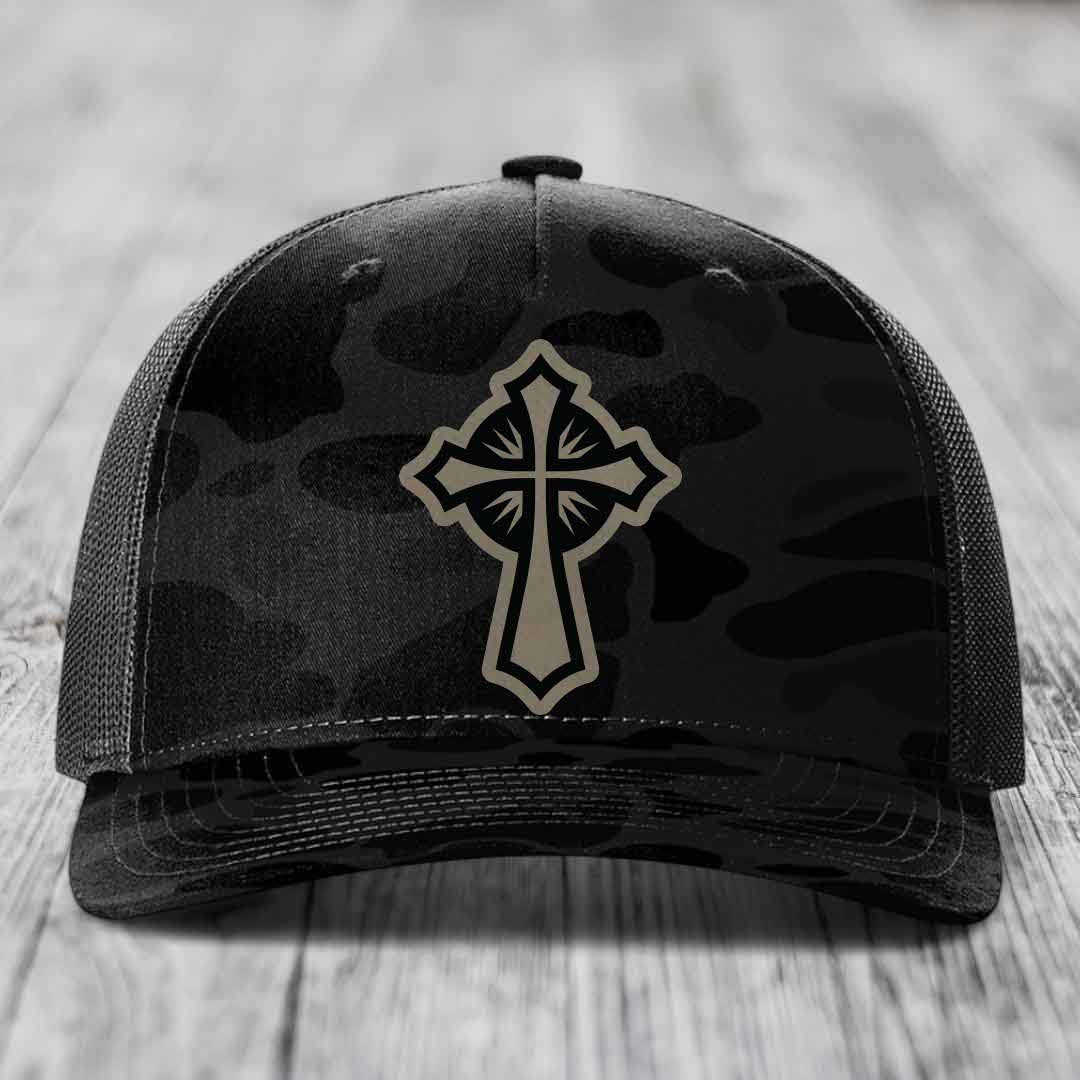 Black camouflage trucker hat with beige cross patch on front, mesh back, sitting on wooden surface.