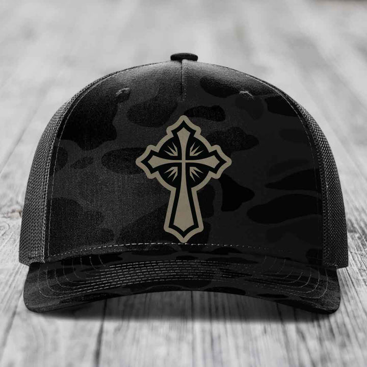 Black camouflage trucker hat with beige cross patch on front, mesh back, sitting on wooden surface.