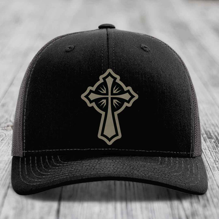 Black trucker hat with a beige cross patch on the front, placed on a wooden surface.
