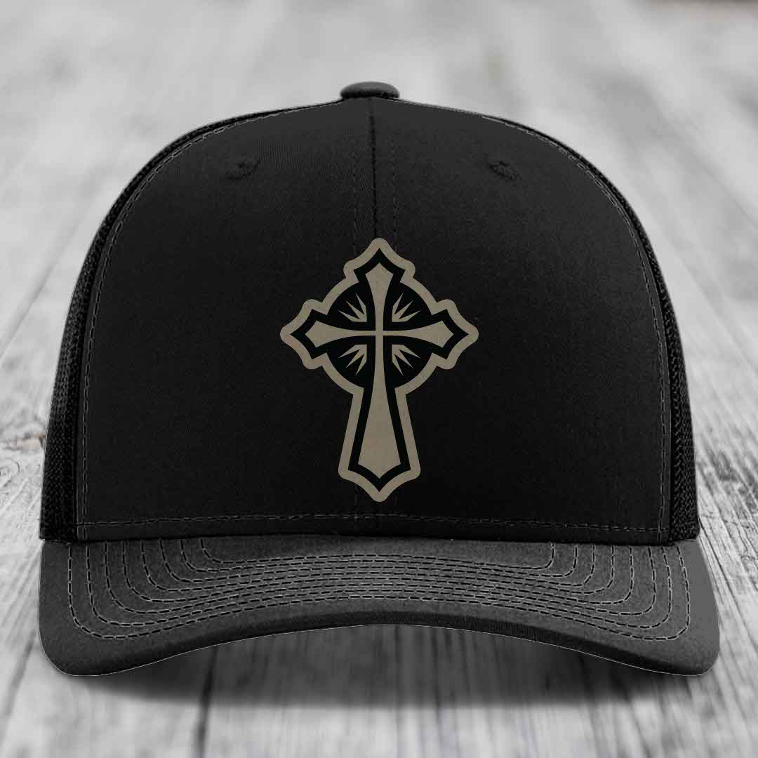 Black trucker hat with a beige cross patch on the front, placed on a wooden surface.
