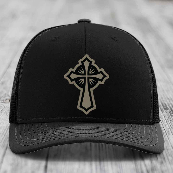 Black trucker hat with a beige cross patch on the front, placed on a wooden surface.