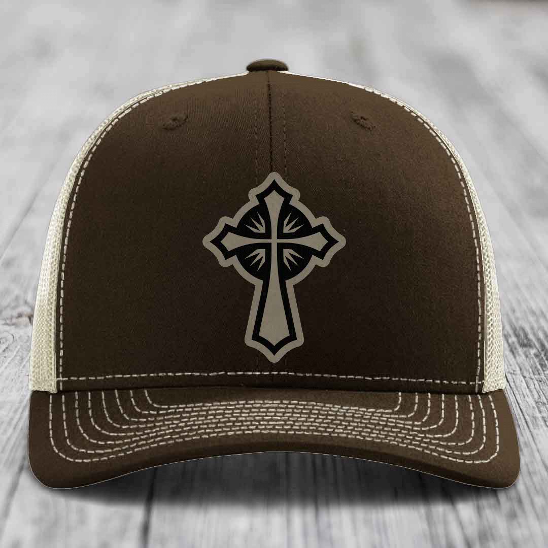 Brown trucker hat with black and beige cross patch, white stitching, and mesh back.