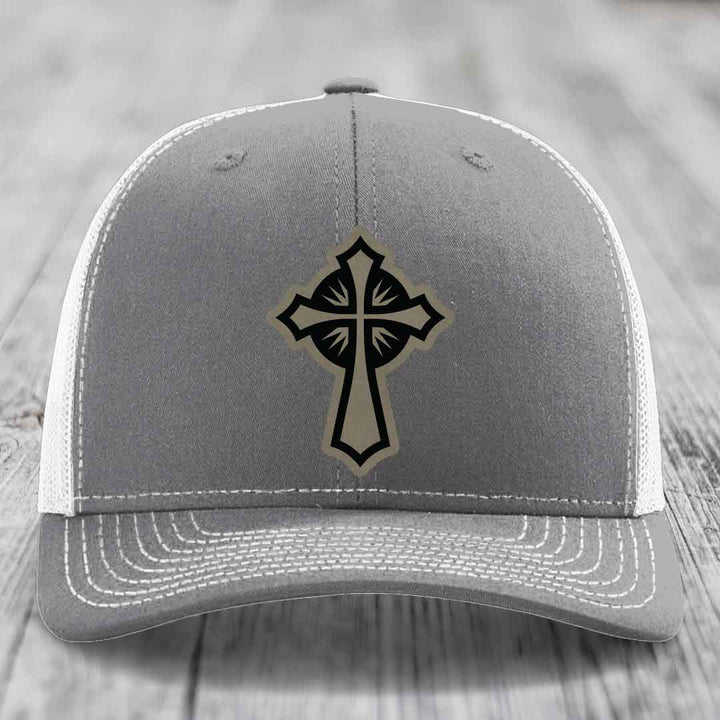 Grey trucker hat with black and tan cross patch on front, white mesh back, white stitching.