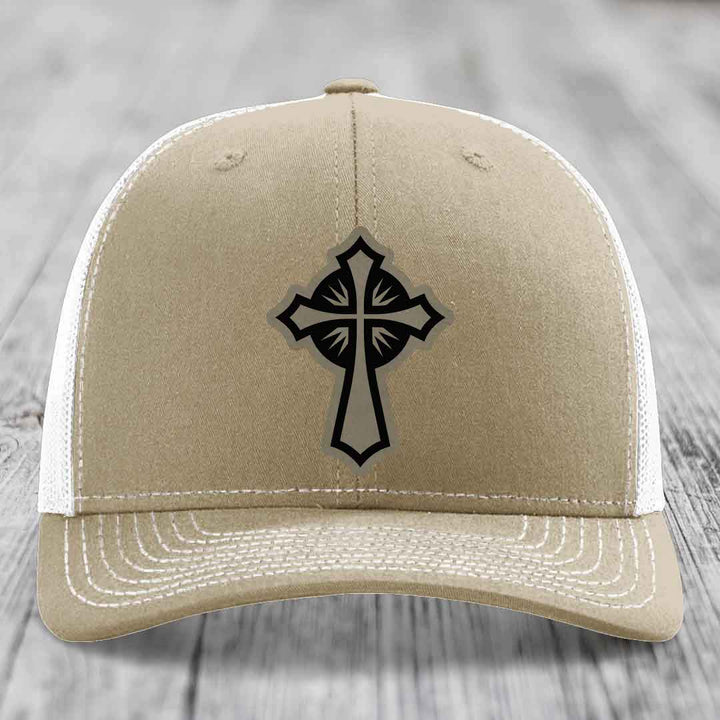 Tan trucker hat with black cross patch on front, white mesh back and white stitching.