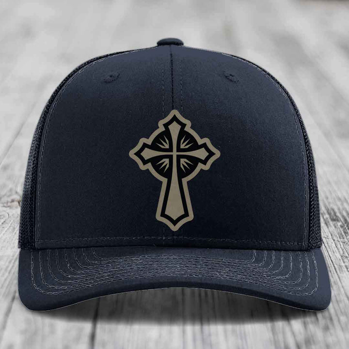Black trucker hat with a beige cross patch on the front, placed on a wooden surface.