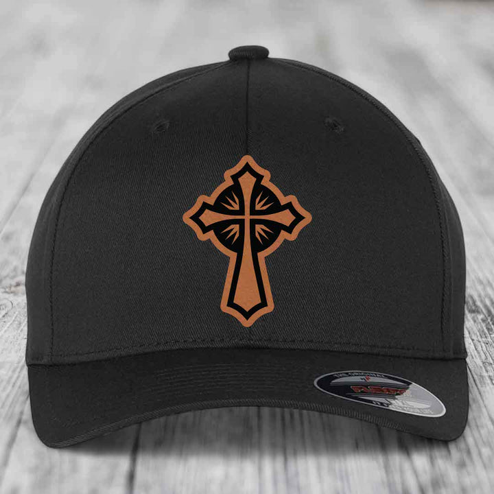Black baseball cap with brown leather cross patch on front, sitting on wooden surface.
