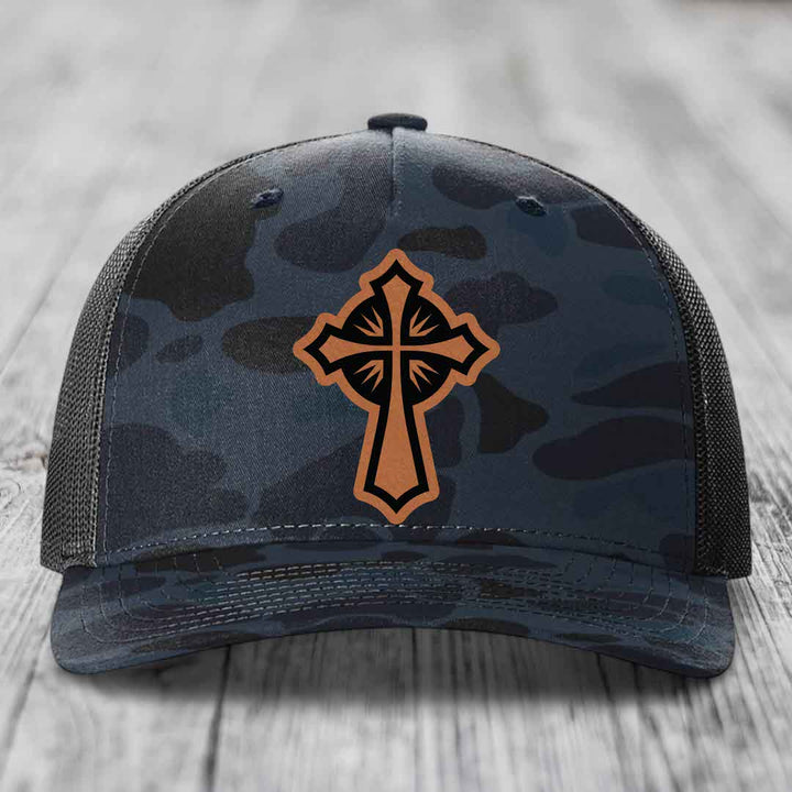 Camouflage trucker hat with brown cross patch on front, black mesh back, resting on wooden surface.