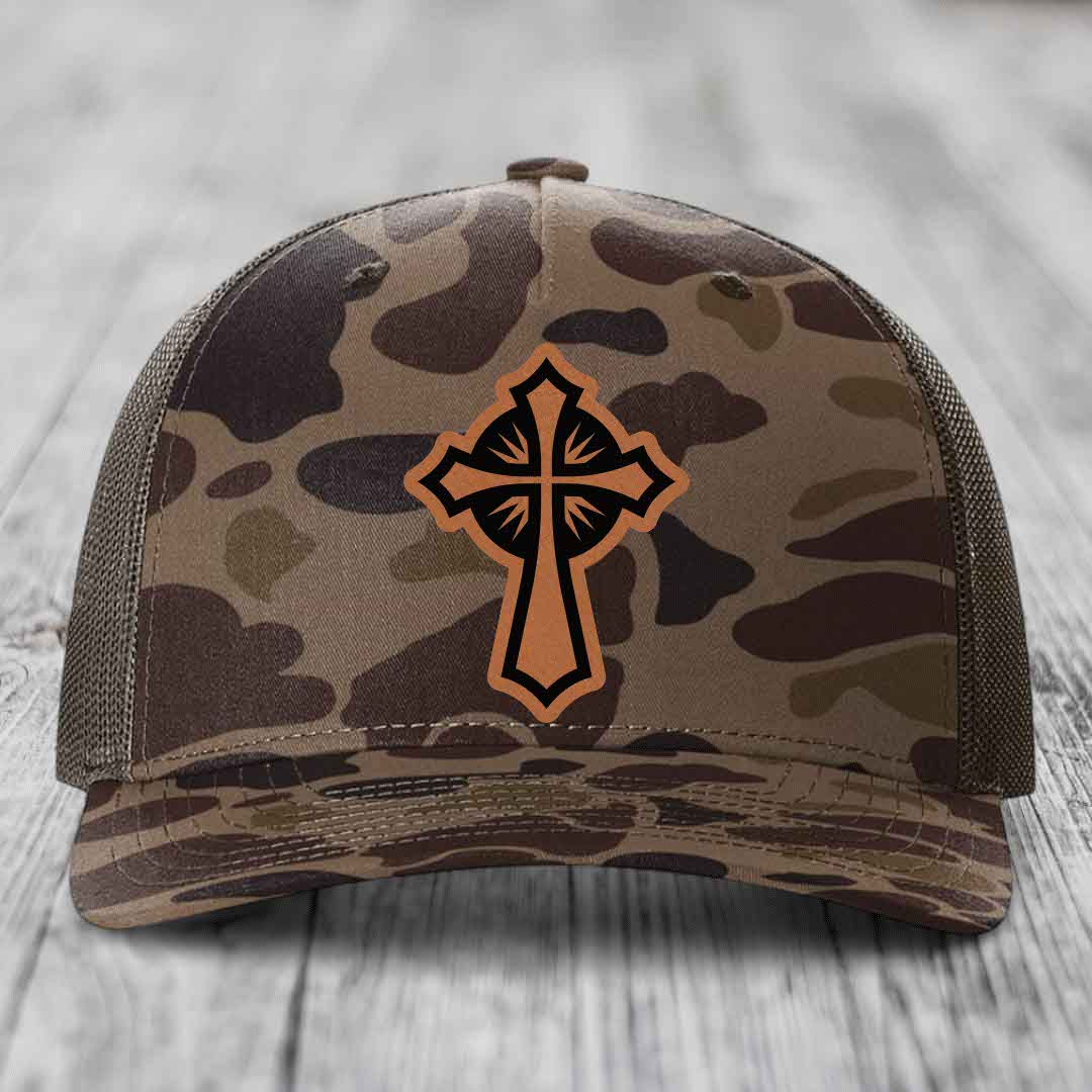 Camouflage trucker hat with brown cross patch on front, mesh back, wooden surface background.