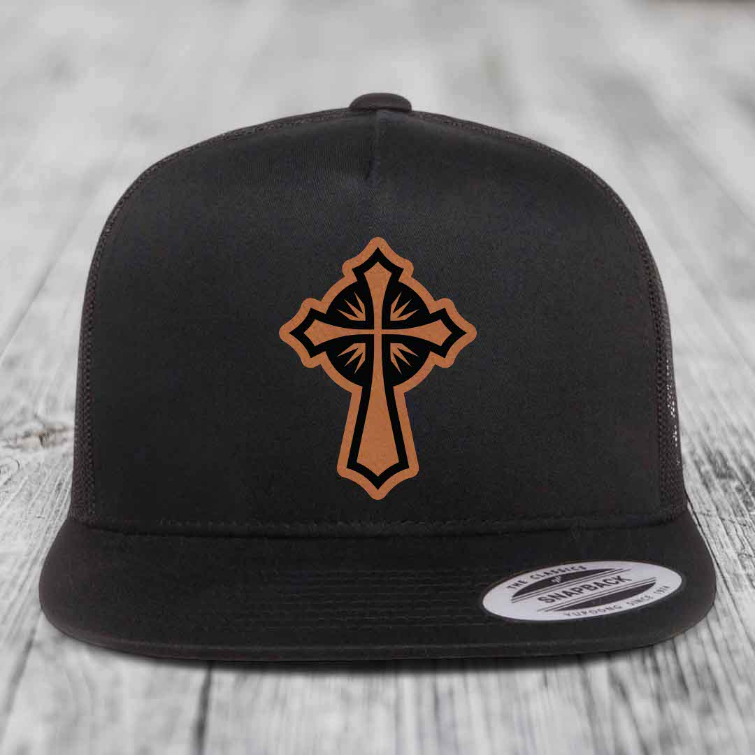Black snapback hat with brown leather cross patch on front, displayed on wooden surface.