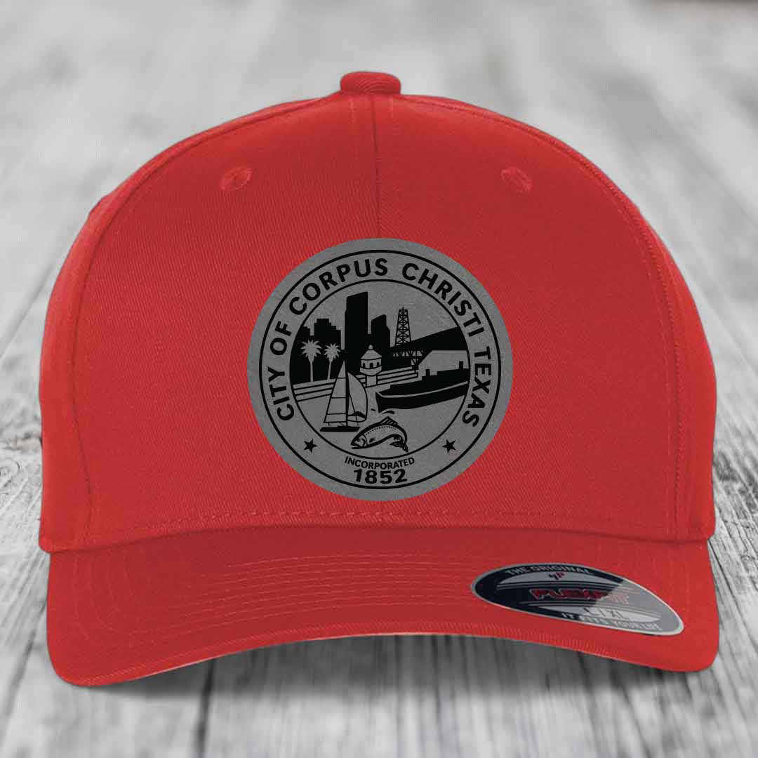 Red baseball cap with grey Corpus Christi Texas city seal patch featuring skyline, boat, and fish.