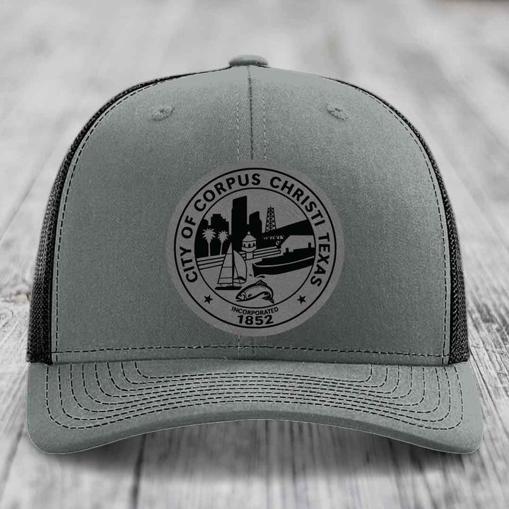 Grey trucker hat with black Corpus Christi Texas city seal patch featuring sailboat, oil rig, and fish.