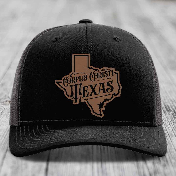 Black trucker hat with brown Corpus Christi Texas leather patch on front, white mesh back and stitching.