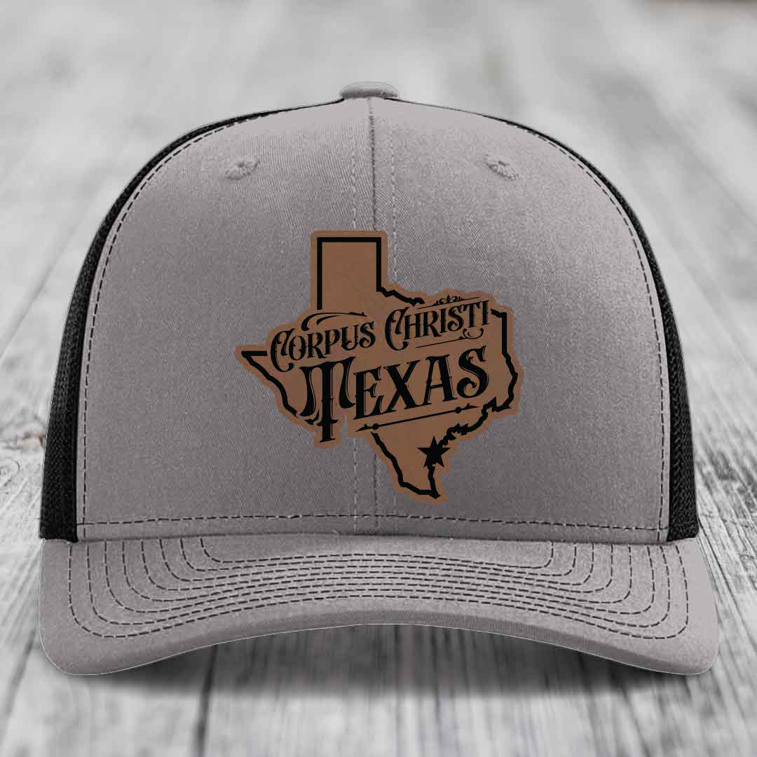 Grey trucker hat with blue mesh sides and a brown leatherette patch shaped like Texas with &