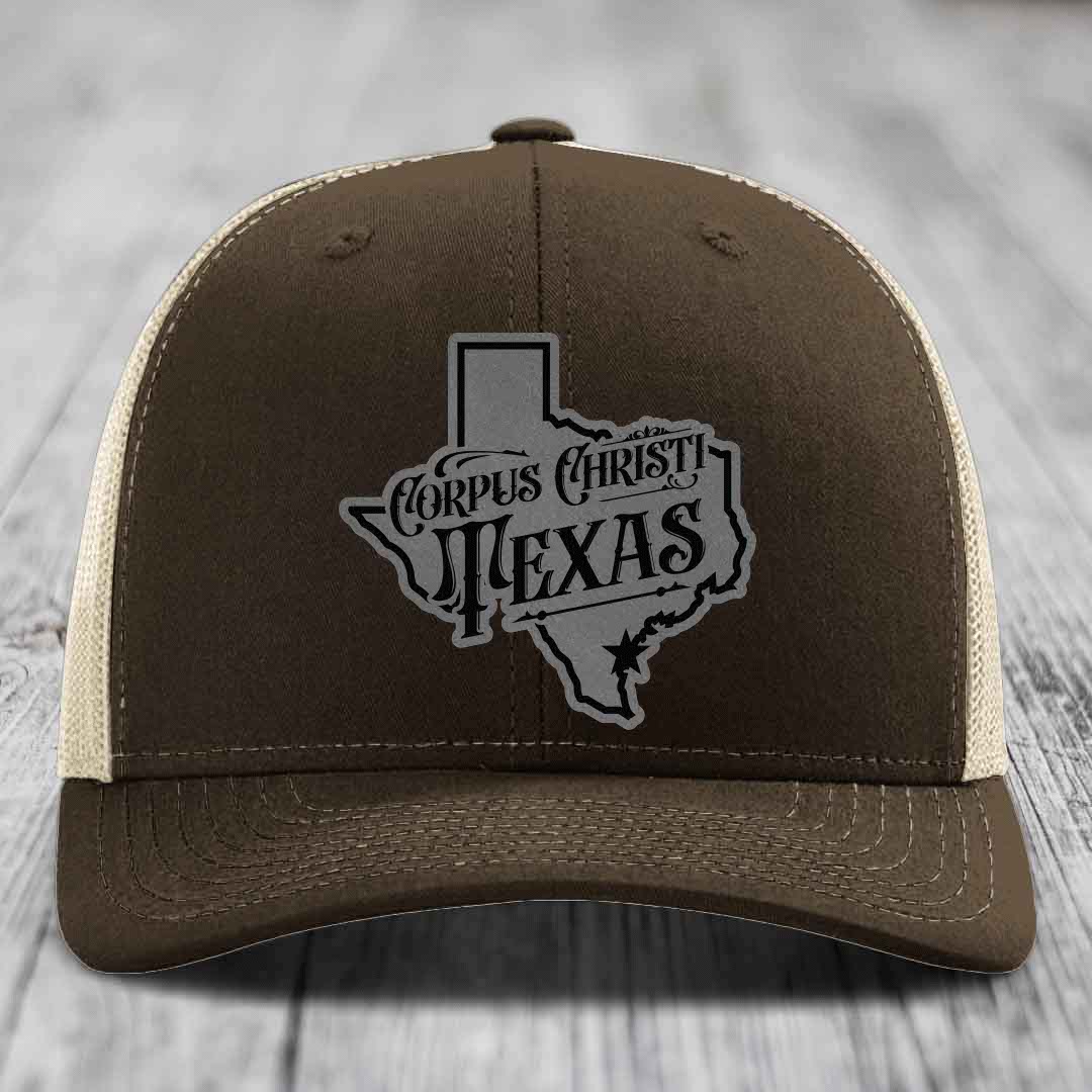 Green trucker hat with gray Corpus Christi Texas patch on front, sitting on wooden surface.
