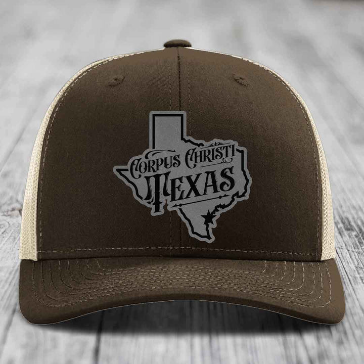 Green trucker hat with gray Corpus Christi Texas patch on front, sitting on wooden surface.
