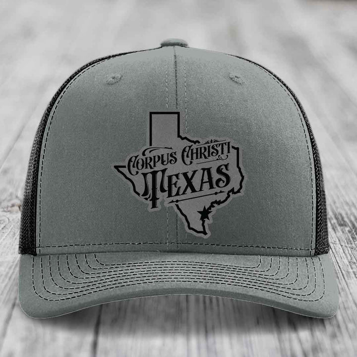 Grey trucker hat with black Corpus Christi Texas patch on front, white mesh back, white stitching.