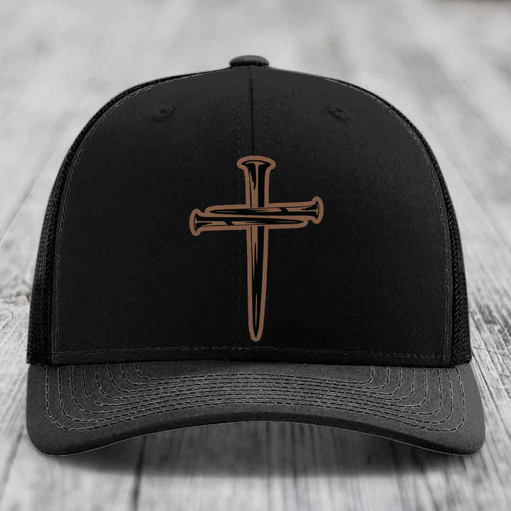Tan baseball cap with blue mesh back and black cross made of nails patch on front.
