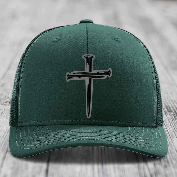 Grey trucker hat with black cross of nails patch on front, placed on wooden surface.